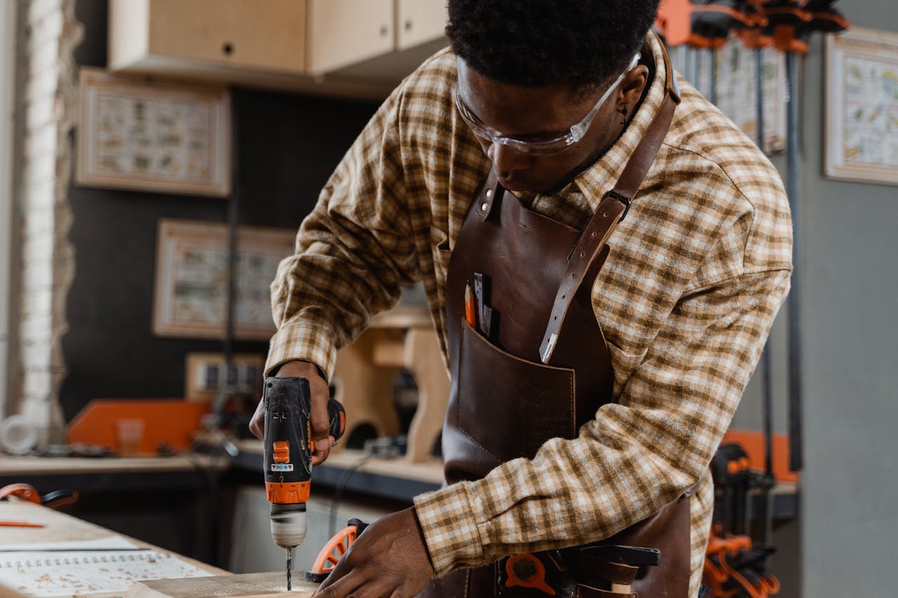 about-02 A craftsman in a workshop focused on woodwork with an electric drill, wearing safety goggles and an apron.
